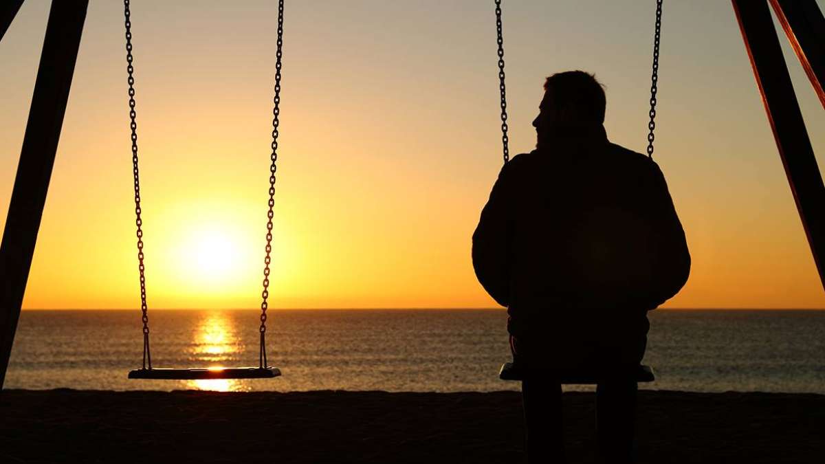 Homem sentado sozinho em um balanço ao pôr do sol à beira do oceano, balanço vazio ao lado dele